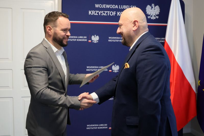W gabinecie Wojewody Lubelskiego stoją naprzeciw siebie i uśmiechając się podają sobie dłonie po lewej stronie doktor Sławomir Kiciak w szarym garniturze po prawej wojewoda Lubelski w granatowym garniturze z żółtą poszetką na tle granatowej ścianki z godłem Polski i napisem „Wojewoda Lubelski Krzysztof Komorski”; 