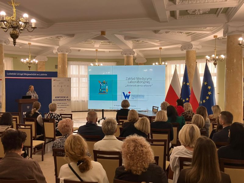 Uczestnicy kikonferencji siedzący na elegancej sali konferencyjnej Lubelskiego Urzędu Wojewódzkiego. Na pierwszym planie widoczne są osoby odwrócone tyłem do obiektywu, podczas gdy prelegentka wygłasza prezentację na mównicy. Na ekranie prezentacji widać tytuł prelekcji dotyczącej Zakładu Medycyny Laboratoryjnej oraz logo szpitala. W tle stoją flagi Polski i Unii Europejskiej oraz banery informacyjne konferencji.
