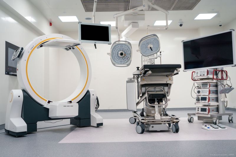 Nowocześnie wyposażona sala operacyjna z dużym, okrągłym ramieniem aparatu obrazującego po lewej stronie, pośrodku z mobilnym stołem operacyjnym i dwiema lampami operacyjnymi podwieszonymi do sufitu oraz po prawej z wysokim monitorem i wózkiem z aparaturą medyczną, w jasnym, sterylnym wnętrzu szpitalnym.