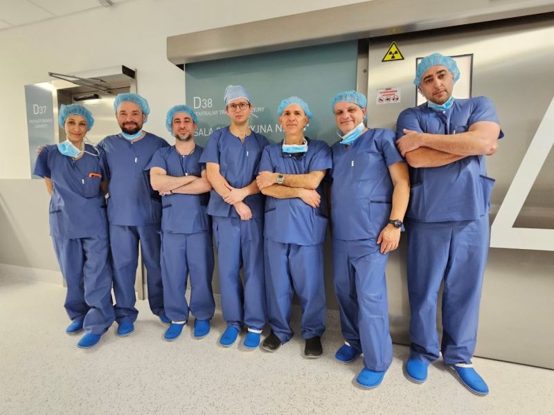 Grupa ośmiu lekarzy w niebieskich uniformach i maseczkach ochronnych, stojących ramię w ramię przed stalowymi drzwiami sali operacyjnej w szpitalu. Tło z tabliczkami „D4” i znakiem ostrzegawczym promieniowania, podłoga w jasnych kafelkach