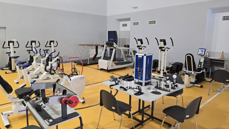 Nowoczesna sala rehabilitacyjna z różnorodnym sprzętem: rowerami stacjonarnymi, bieżnią, orbitrekami, stołem do masażu, maszynami do ćwiczeń nóg i rąk oraz stołem z akcesoriami. Jasne pomieszczenie z niebieskimi matami i drewnianą podłogą