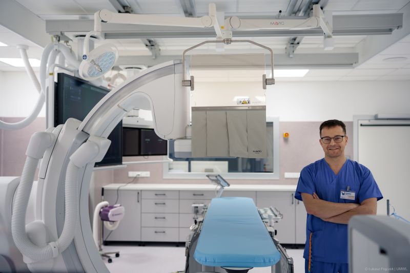 Kierownik zakładu Radiologii dr Maciej Mazgaj w niebieskim uniformie medycznym z identyfikatorem na piersi stoi z założonymi rękami i uśmiecha się do aparatu w jasnej sali radiologii zabiegowej szpitala. W tle widoczny jest zaawansowany angiograf z ruchomym ramieniem C, monitory wyświetlające obrazy medyczne, stół zabiegowy z niebieską matą oraz wyposażenie sali - lampy, szafki i panele sterujące.