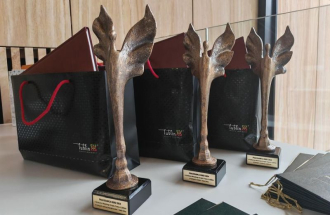 Na jasnym stole ustawione są trzy metalowe statuetki z figurą stylizowanego anioła o uniesionych skrzydłach, umieszczone na czarnych postumentach z napisem „Pracodawca Roku 2024”, a za nimi stoją czarne torby prezentowe z czerwonymi uchwytami i logo „Lublin – Miasto Inspiracji”; obok widoczne są eleganckie teczki i dyplomy przygotowane na uroczystą galę wręczenia nagród.