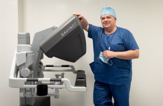Na jasnym tle sali operacyjnej stoi uśmiechnięty Dr Jerzy Draus w niebieskim stroju operacyjnym i czepku, opierając rękę na dużej szarej konsoli robota chirurgicznego da Vinci