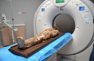 Zdjęcie przedstawia egipską mumię dziecka umieszczoną na stole badawczym wewnątrz nowoczesnego tomografu komputerowego